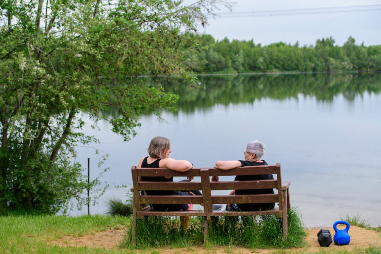 Two ladies in workout clothes sat on a bench by a lake