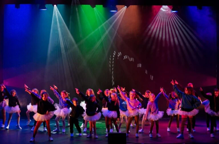 A group of children many in tutus dance on stage with green, red and blue lights in the background.