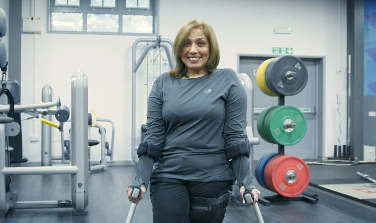 A women in grey top with crutches stand in front of gym equipment.