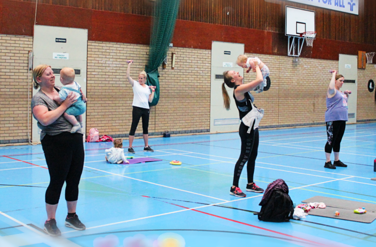 A group of new mums and pregnant women with babies taking part in a workout in a sports hall.