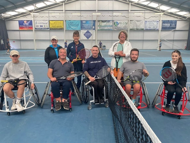 A group of people, some standing and some in wheelchairs pose for a photo on an indoor tennis court.