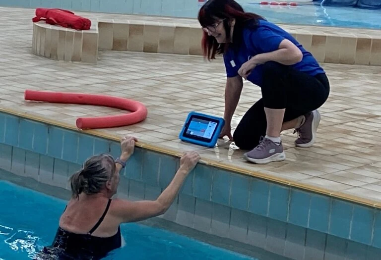 A women in a pool talks to the instructor at the side, whilst looking at the poolside tablet.