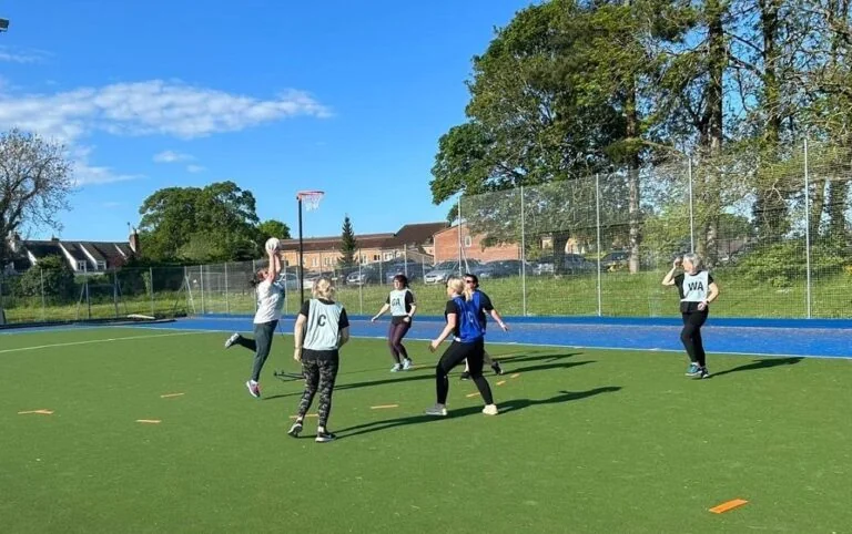 A group of women in blue netball bibs, playing netball on an outdoor court. One lady is jumping as she catches the ball.
