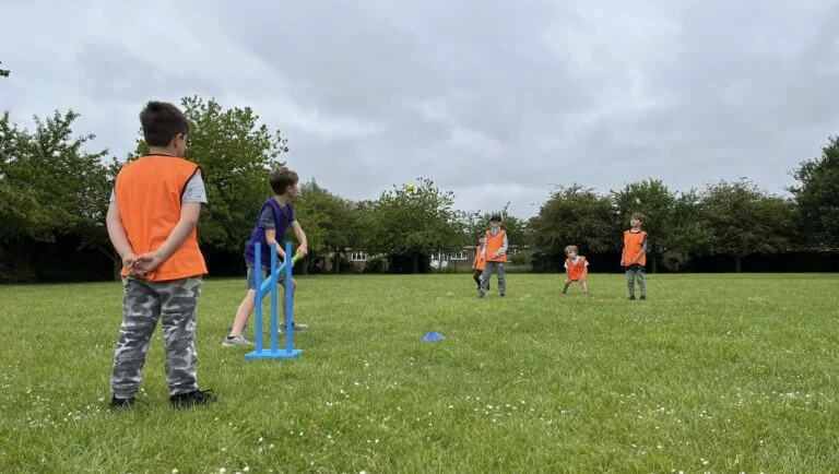 A group of children in orange hi-vi vests play cricket on a grass playing field
