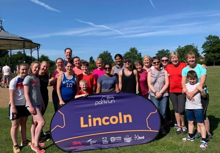 A group of people in running clothes pose for a photo on a summer day in front of purple parkrun pop up banner.