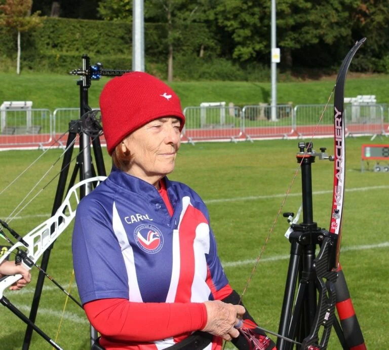 A partially sighted women (Carol) wearing a red and blue kit, holds a archery bow.