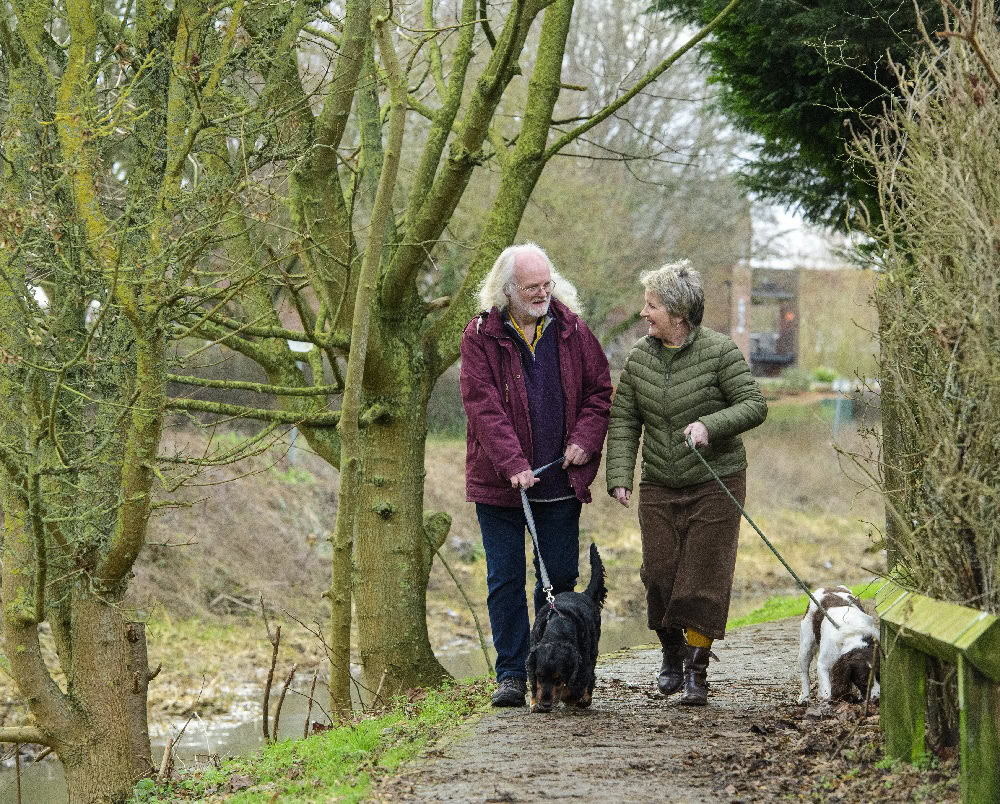 A man and a women in winter clothing, take a dog for a walk down a tree lined path.