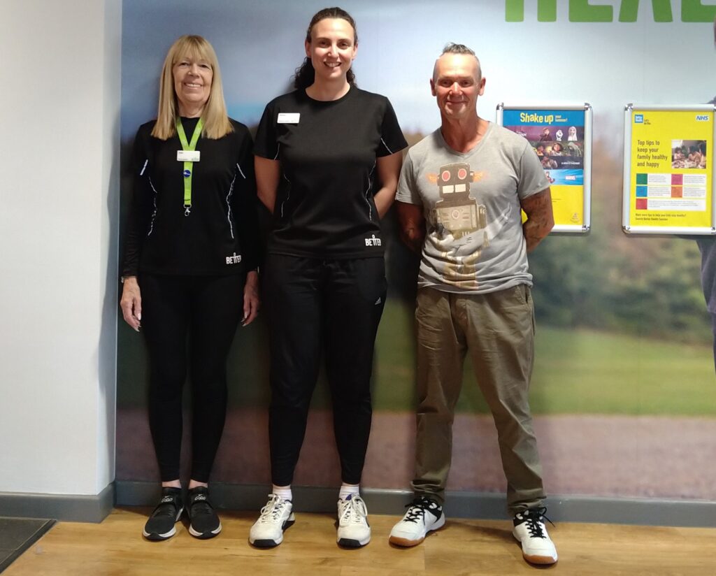 Two women in fitness clothes stand next to Tommy and pose for a photo, against a backdrop of a leisure centre wall with posters.