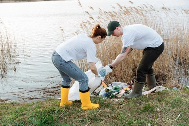 A man and a woman in white t-shirts and wellington boots, put litter next to a lake into a bag.