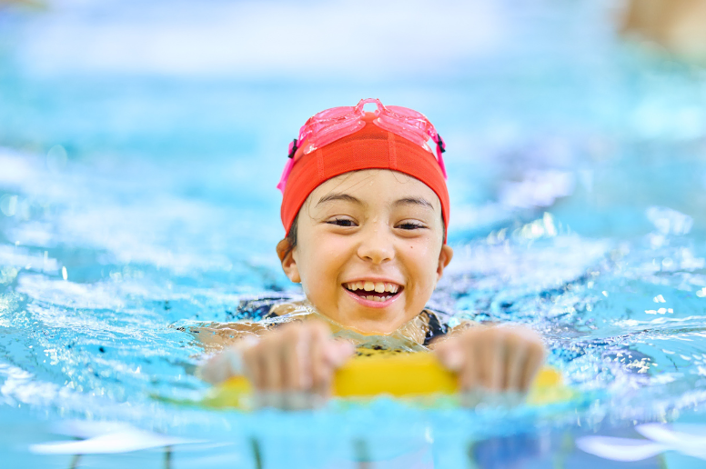 A smiling girl in a red swimming cap, swims in  a pool using a float.