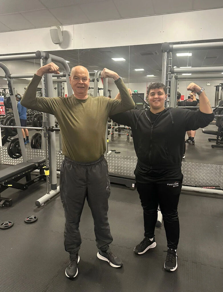 An older man poses next to his personal trainer in the gym, they pose for a photo with arms flexed.