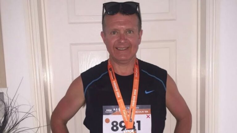 A man in running vest, with a number on and lanyard poses for a photo inside a house.