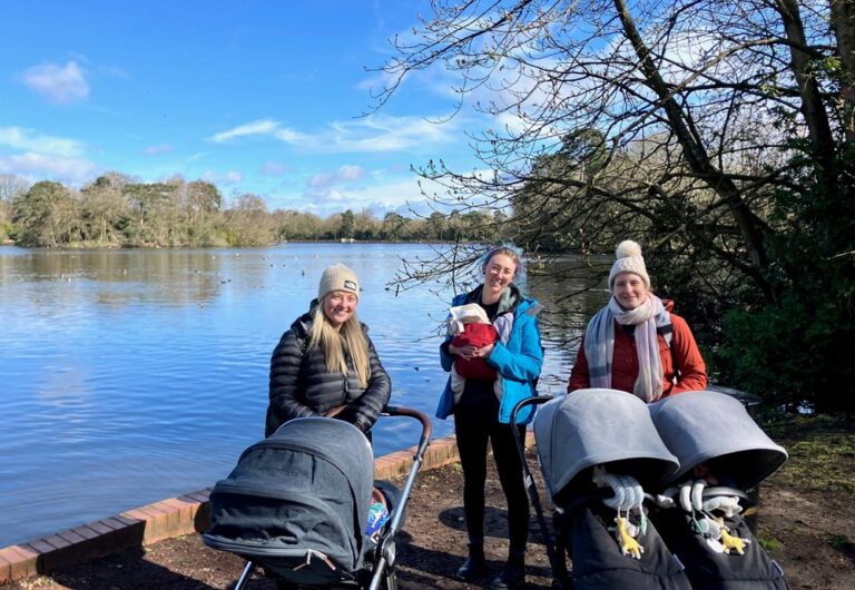 A group of mums,with babies in arms or buggies, pose in front of a lake, with a blue sky and trees.